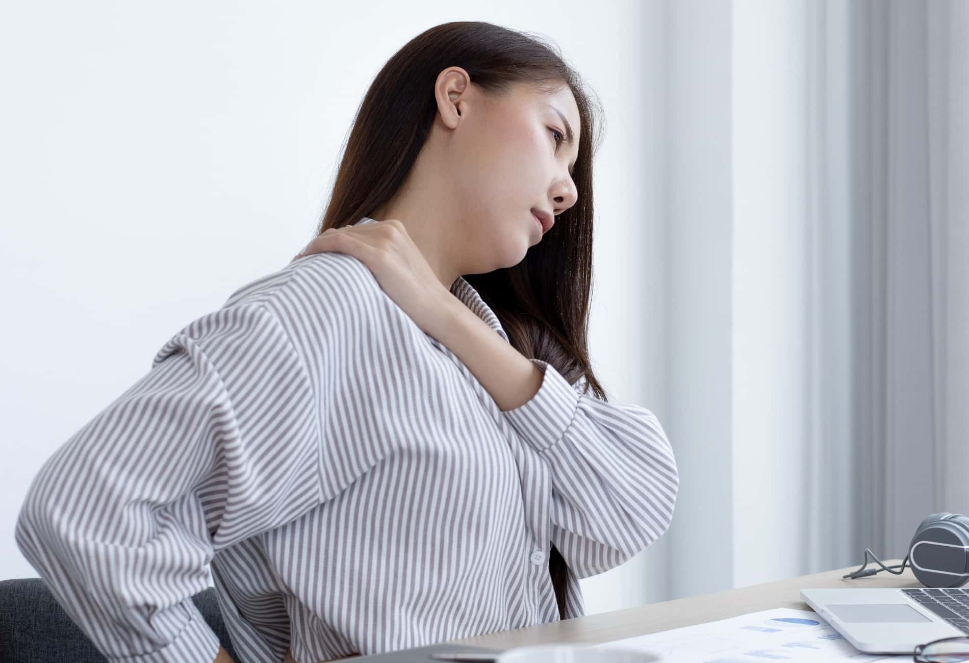 Mulher sentada em sua mesa de escritório, segurando o pescoço e a nuca com as mãos. A imagem representa os sintomas de hérnia de disco cervical causados por má postura e o impacto da dor no dia a dia de trabalho.