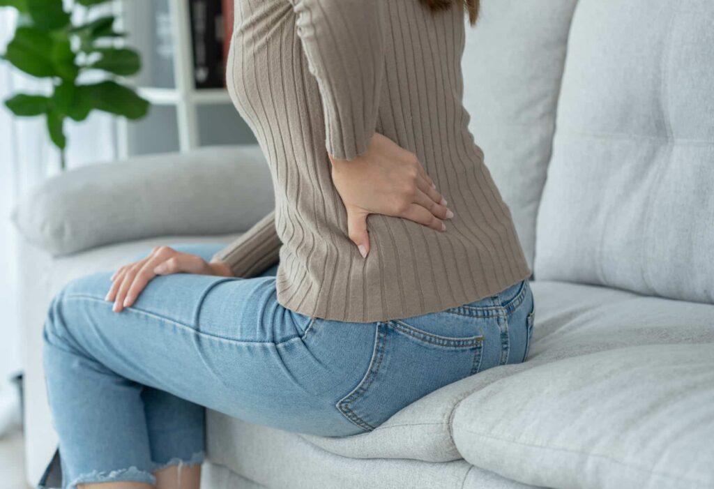 Mulher sentada em um sofá, com a mão na parte inferior das costas, sentindo dor lombar. A imagem representa o desconforto diário e os sintomas causados pela hérnia de disco, destacando a importância de buscar tratamento.