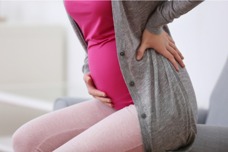 Mulher grávida sentada, segurando a região lombar e a barriga com as mãos, indicando dor nas costas. A imagem representa o aumento de risco para hérnia de disco durante a gestação e a importância de buscar um tratamento não cirúrgico e seguro.