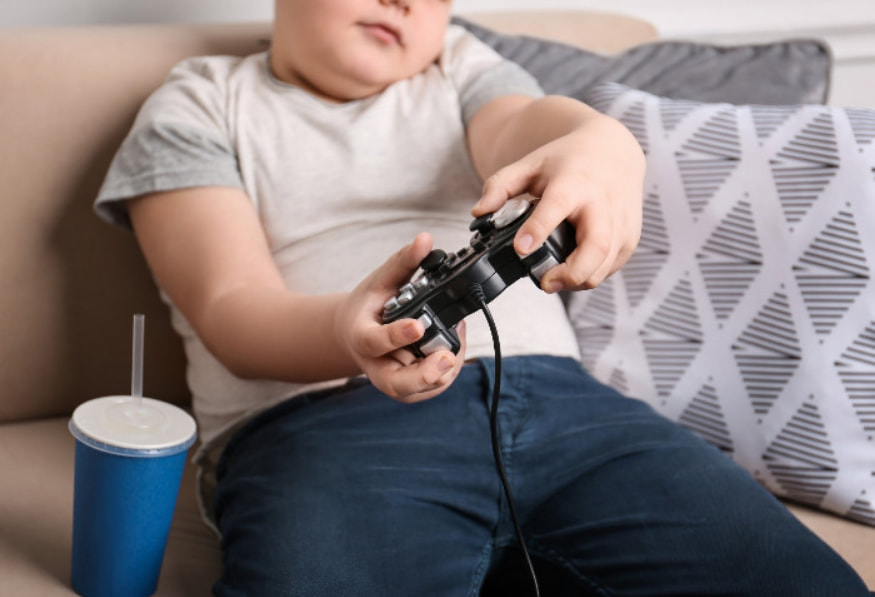 Criança obesa sentada no sofá com um controle de videogame nas mãos, ao lado de um copo de refrigerante. A imagem ilustra o sedentarismo e a má postura, fatores de risco que contribuem para o desenvolvimento de uma hérnia de disco em jovens.