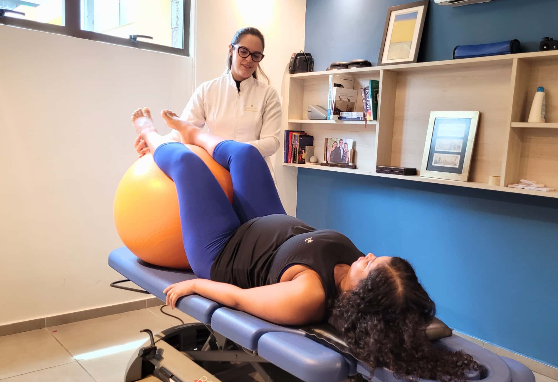 Fisioterapeuta auxiliando paciente a realizar um exercício com bola suíça, deitada em uma maca. A imagem mostra o tratamento não cirúrgico e a fisioterapia para hérnia de disco, focando no fortalecimento muscular para alívio da dor.