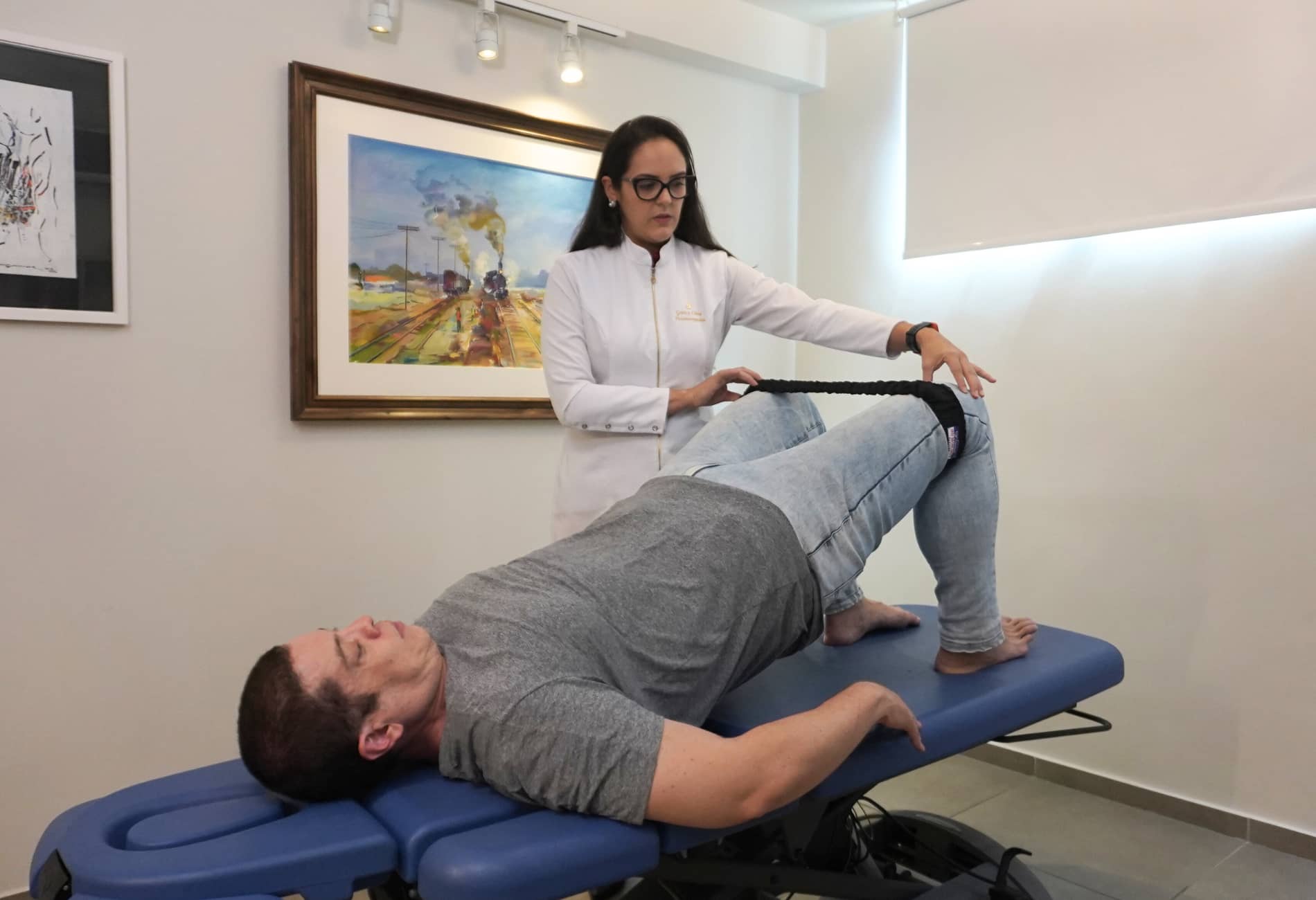 Fisioterapeuta conduzindo exercício para dor lombar, onde o paciente realiza a ponte de glúteos com faixa elástica para intensificar o fortalecimento.