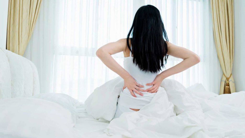 mulher sentada na cama com as duas mãos na lombar
