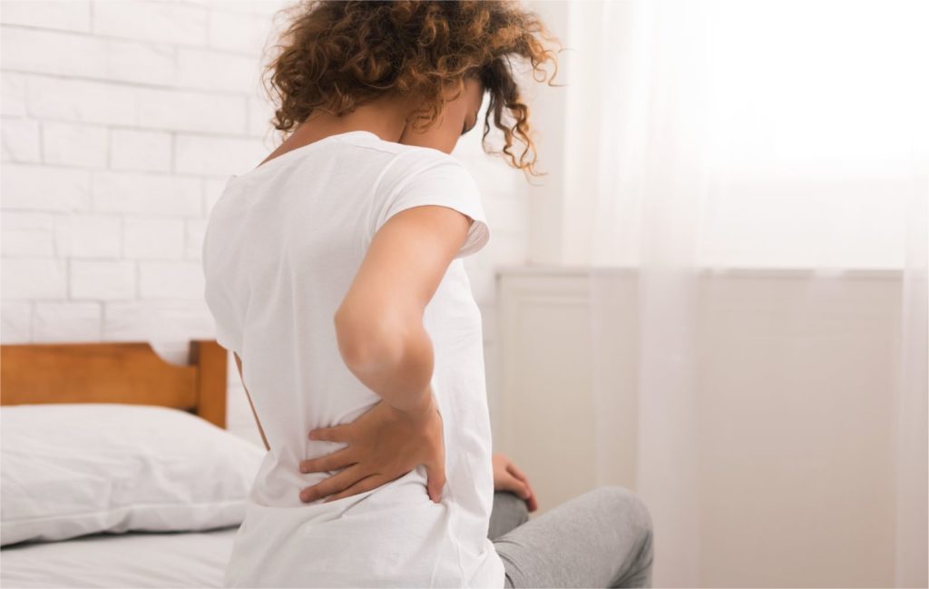mulher sentada na cama com uma das mãos nas costas sentindo dor no cisto de tarlov
