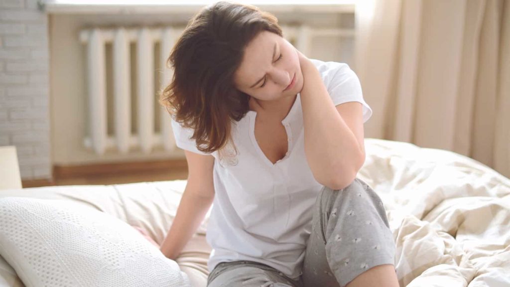 mulher sentada na cama com uma das mãos no pescoço ocm dor por síndrome do chicote.