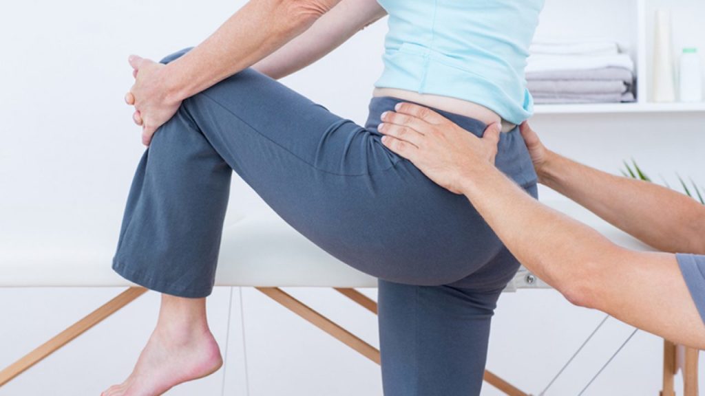 mulher de costas com uma das pernas levantadasfazendo Alongamento Piriforme com profissional dando suporte