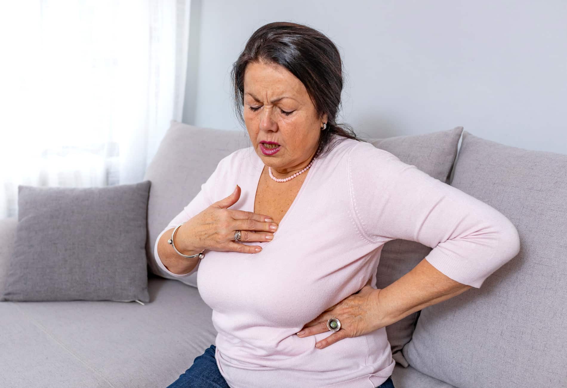Mulher mais velha sentada no sofá com expressão de desconforto, segurando o peito com a mão direita e a região lombar com a mão esquerda, simbolizando a dor no peito e nas costas.