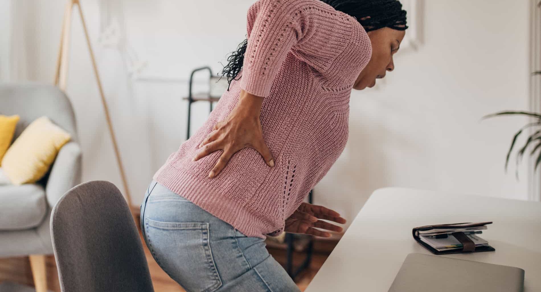 Uma mulher de suéter rosa sentindo dor lombar ao tentar se levantar de uma mesa de escritório.